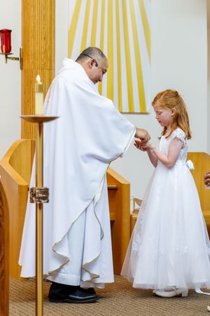 A priest in white robes performs a religious ceremony with a young girl wearing a white communion dress in a church.