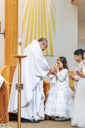 A Catholic First Communion ceremony shows children receiving Holy Eucharist from a priest in white robes at the altar.