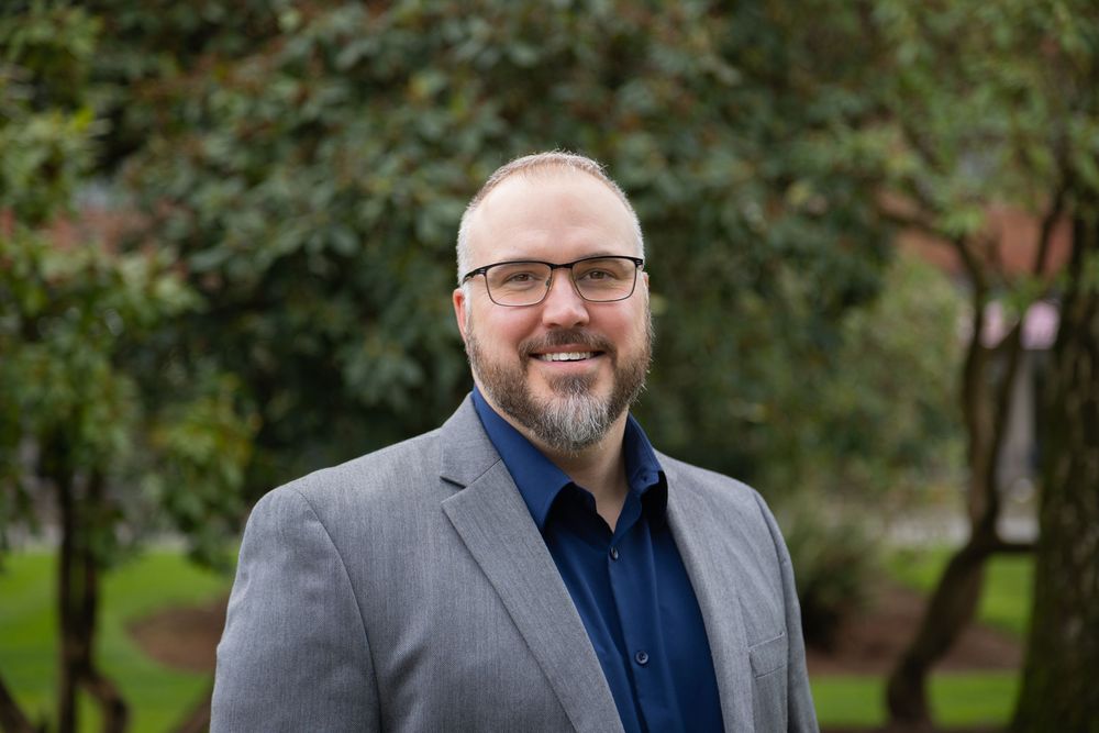 Professional man with beard and glasses wearing gray blazer smiling outdoors in a green garden setting.
