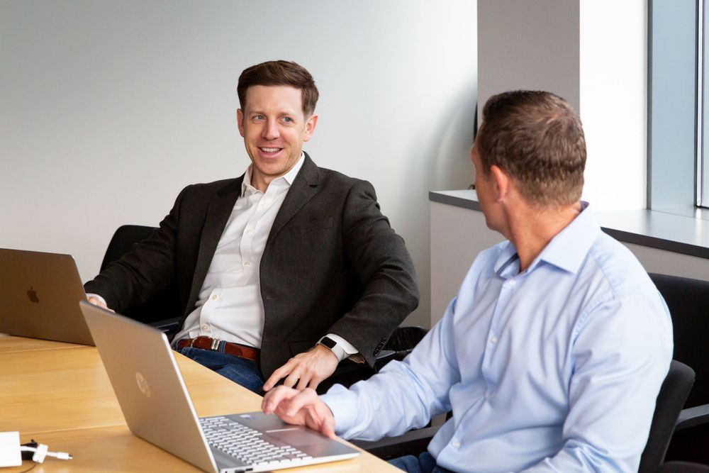 Two professionals having a business discussion while working on laptops in a bright office space.