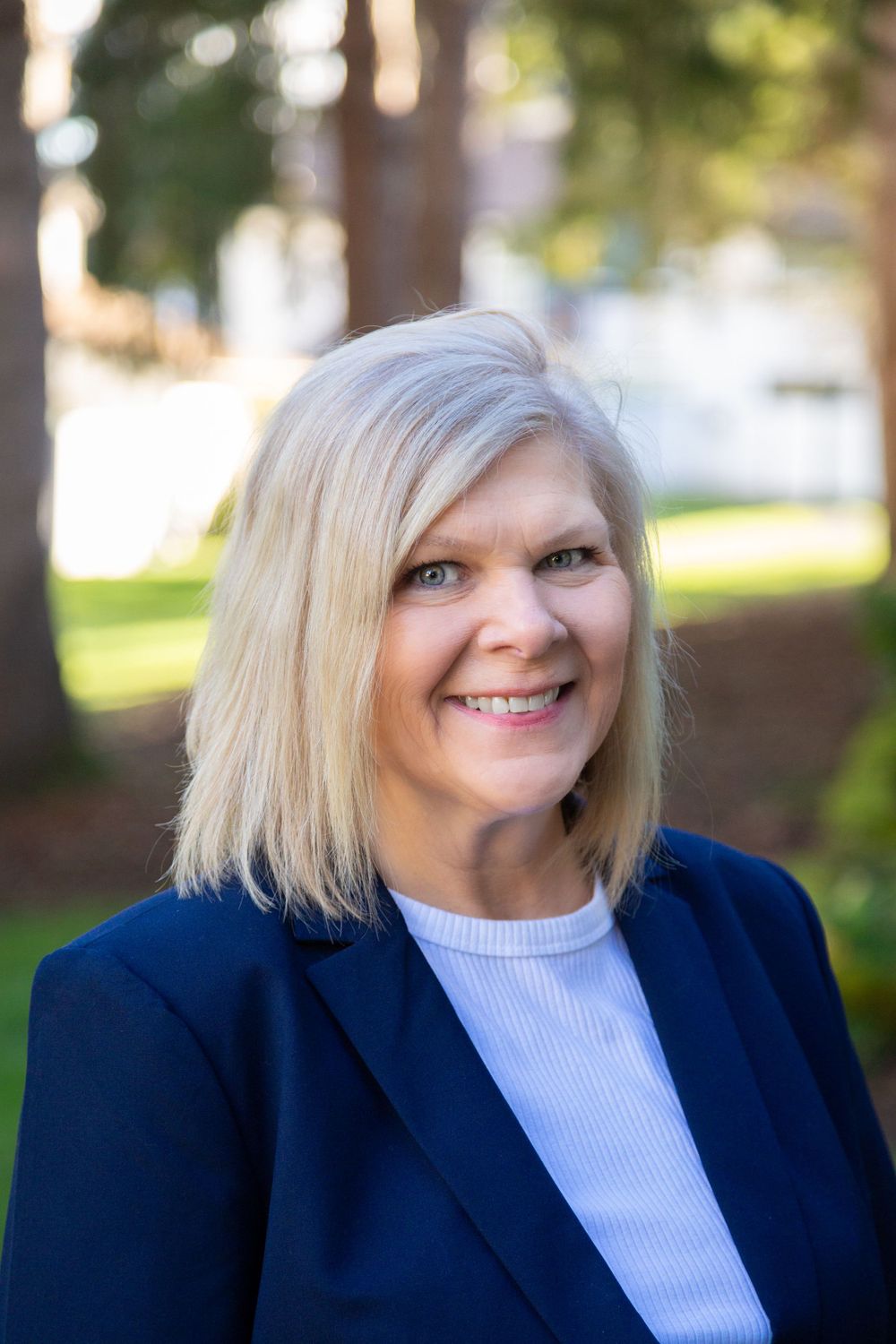 Professional headshot of someone wearing a navy blazer and white top against a blurred outdoor background.