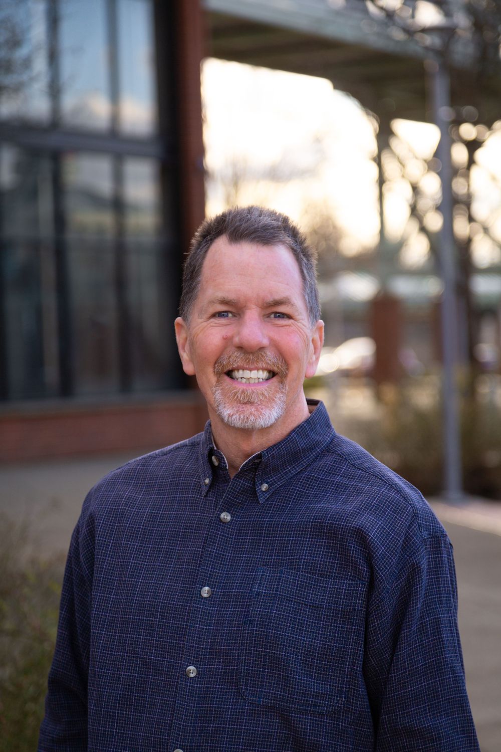 Professional headshot of a person wearing a navy button-down shirt standing outdoors with a warm sunset background.