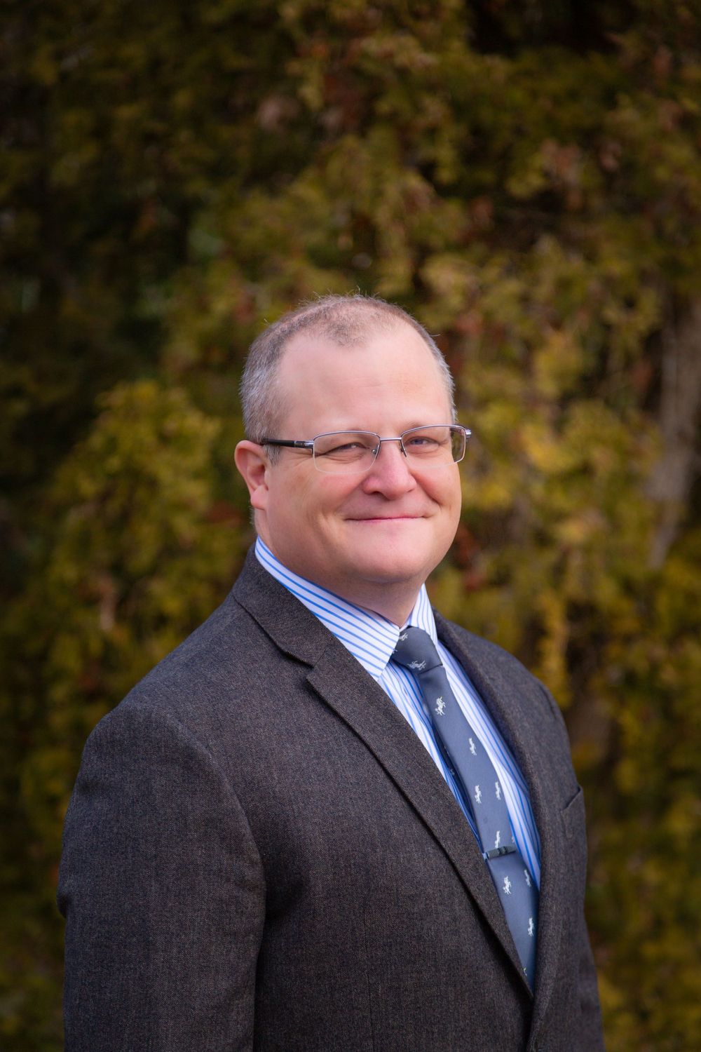 Professional headshot of a person in a dark suit jacket, striped shirt and blue tie against an autumn background.