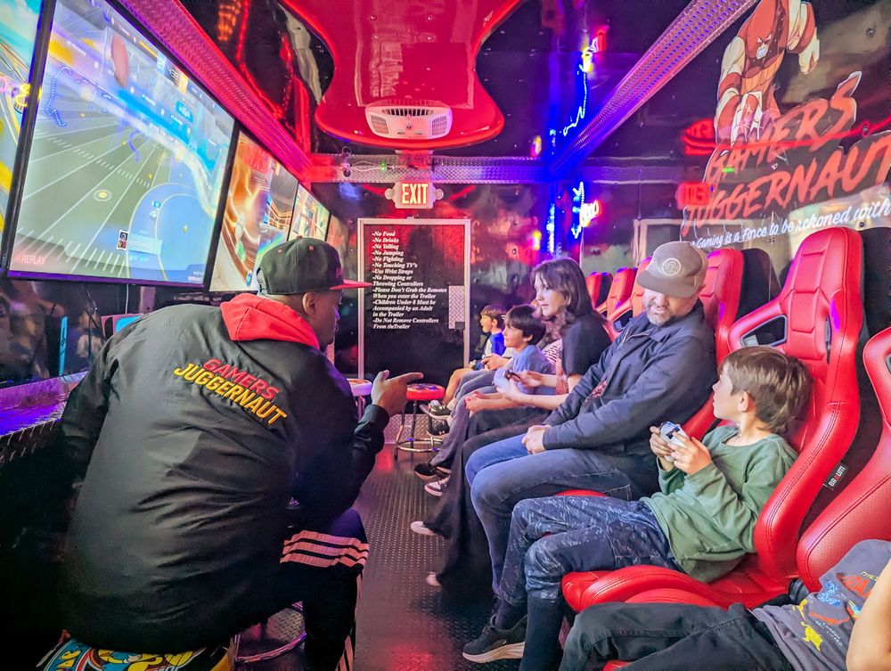 People sit in red gaming chairs at an arcade with colorful neon lighting and multiple screens visible.