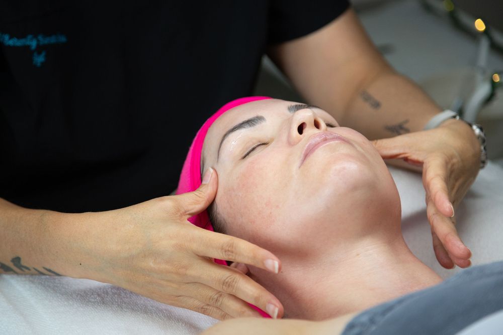 A woman with pink hair receives a relaxing facial massage treatment at a beauty salon from a tattooed therapist.
