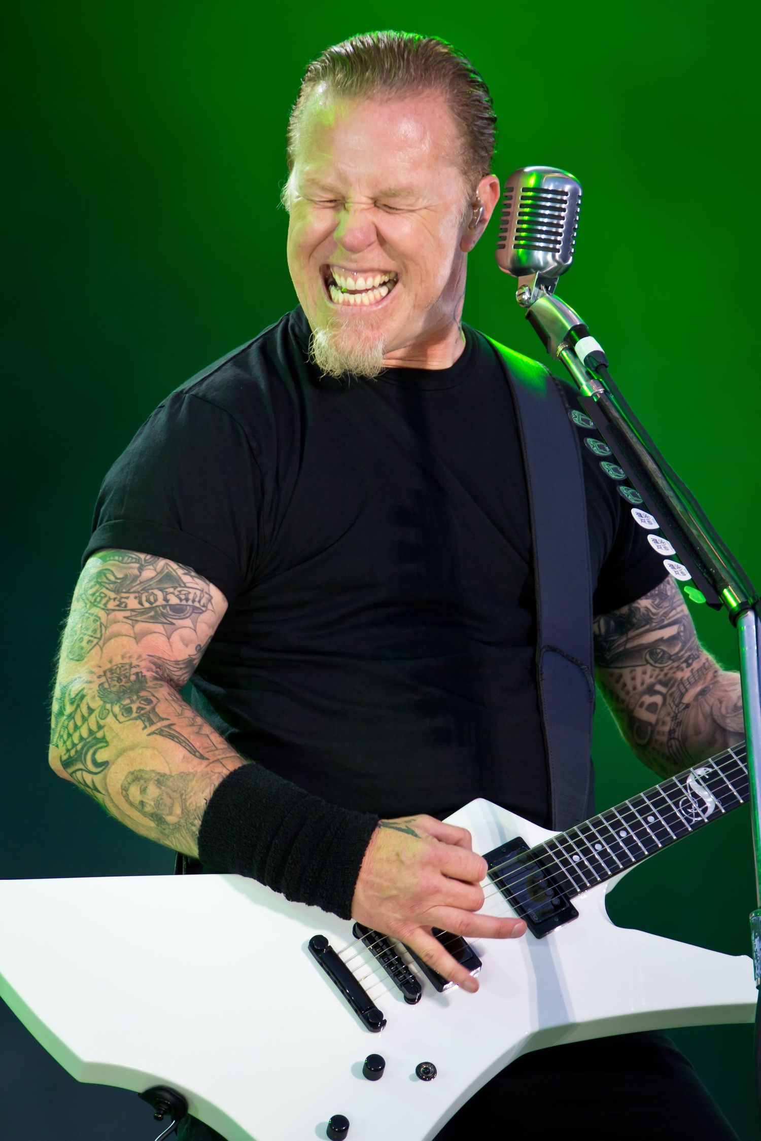 Tattooed rock guitarist smiling while playing white electric guitar on stage with green lighting.