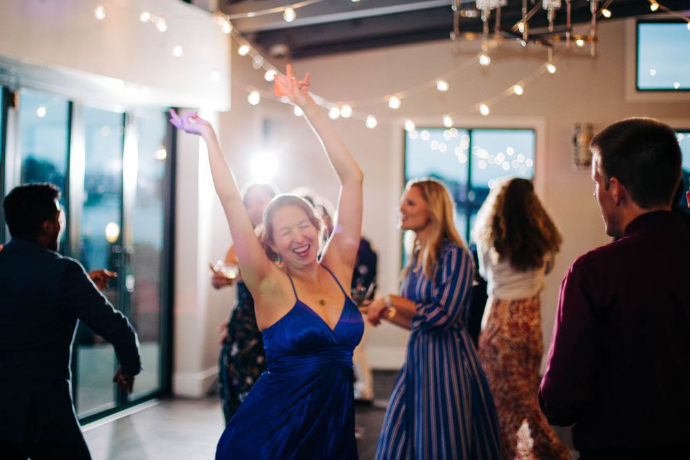 Guest in blue dress dances enthusiastically under string lights at wedding reception.