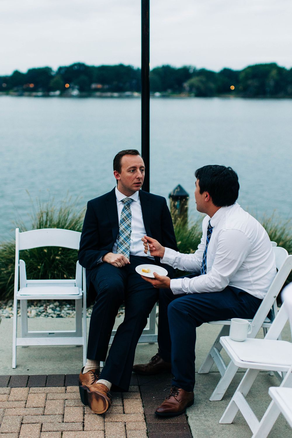 Two businessmen having a casual conversation while seated on white chairs by a waterfront at dusk.