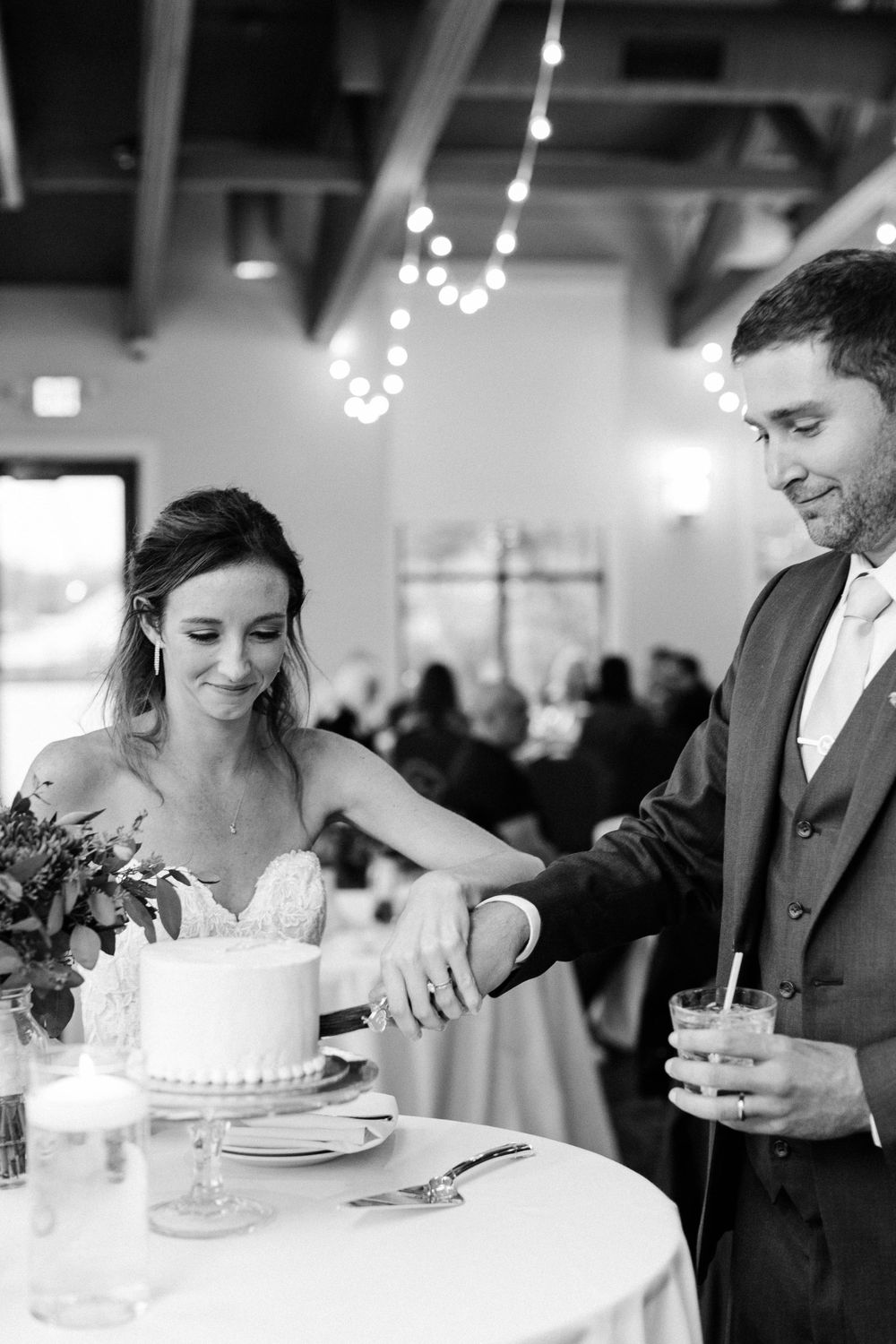 Newlyweds cut their elegant white wedding cake together at reception with string lights overhead.