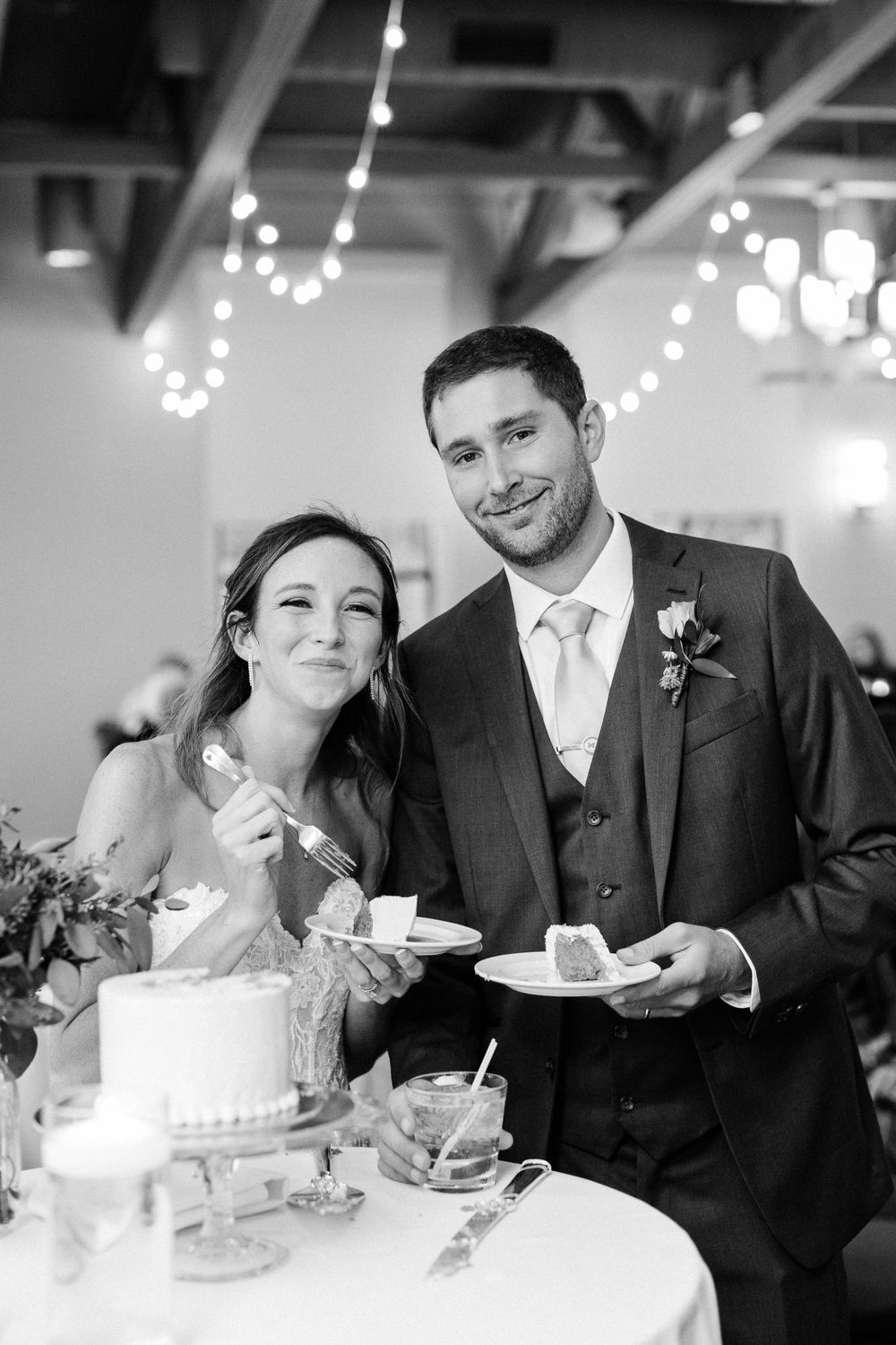 Newlyweds share cake at reception while standing together under string lights.