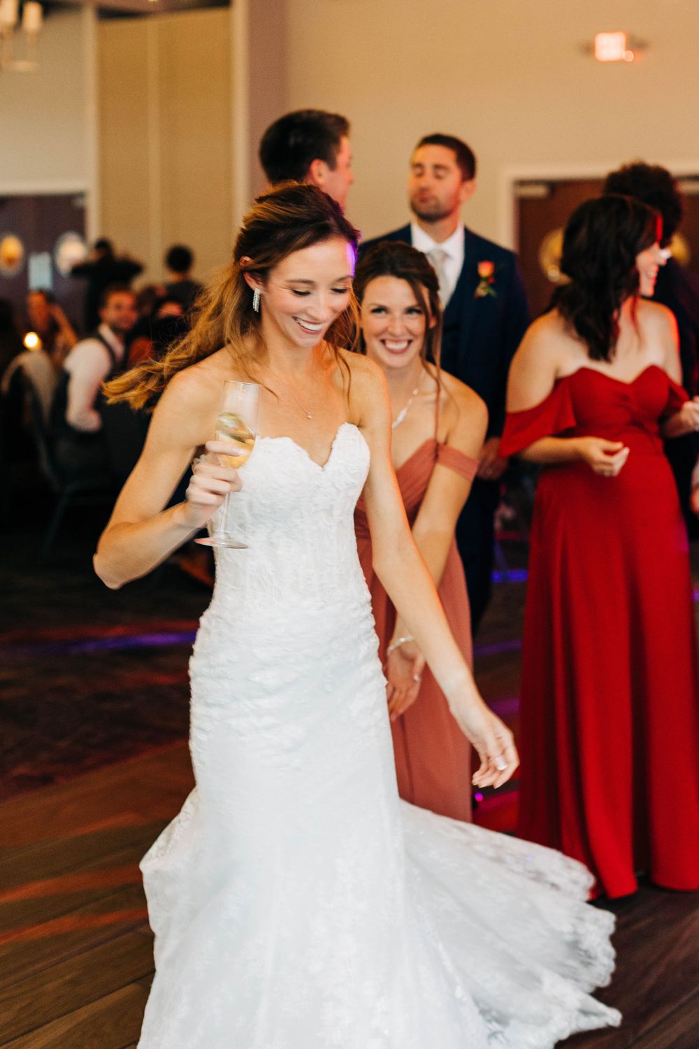 Bride in white lace gown dances joyfully at wedding reception with guests in background.