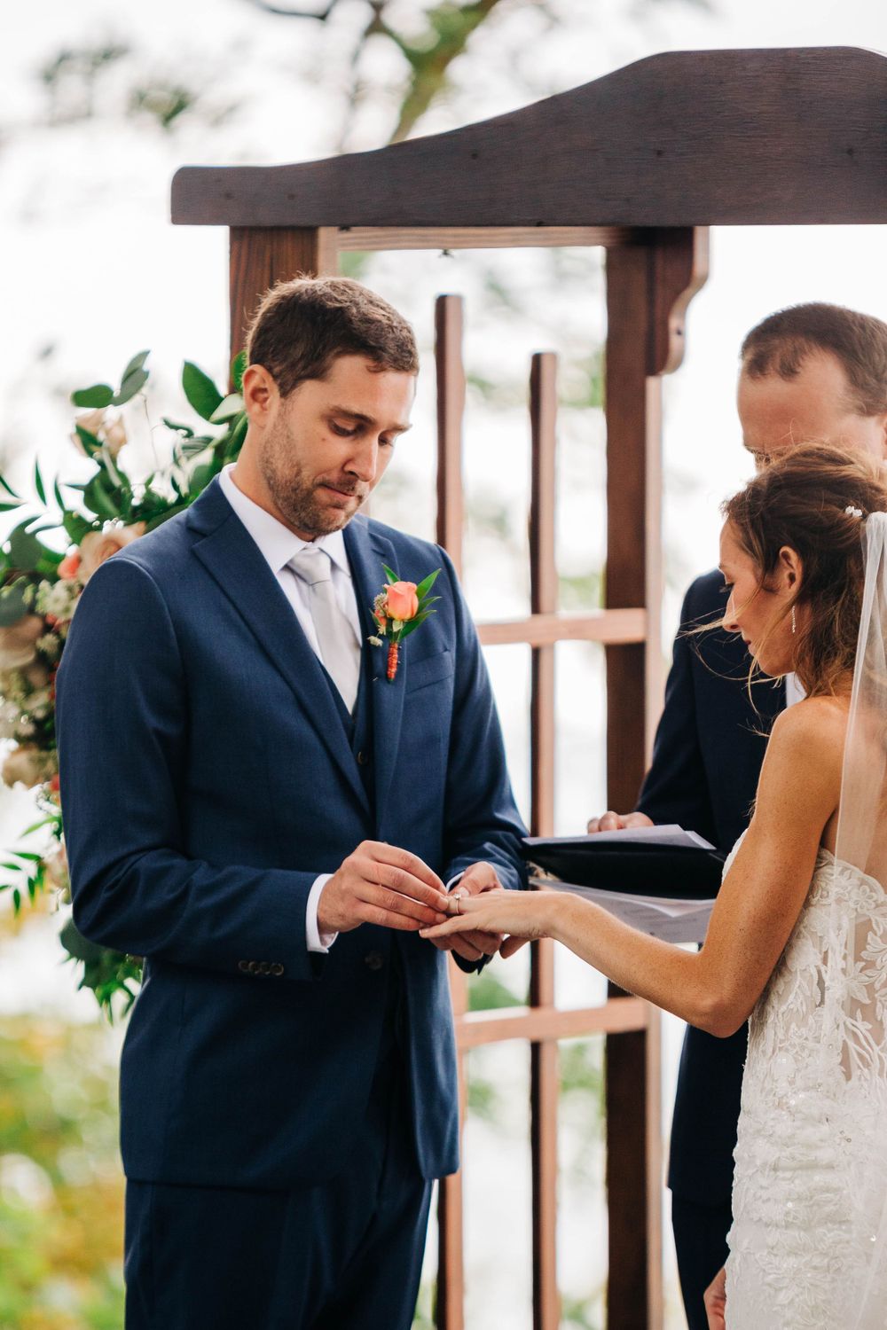 A couple exchanges rings during their wedding ceremony under a wooden arch decorated with greenery.