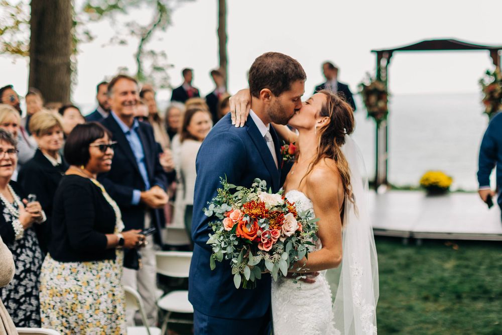 Newlyweds share a celebratory kiss while guests applaud during outdoor ceremony.
