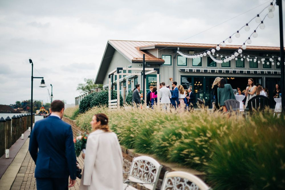 Wedding guests gather outside a waterfront venue with string lights and decorative touches.