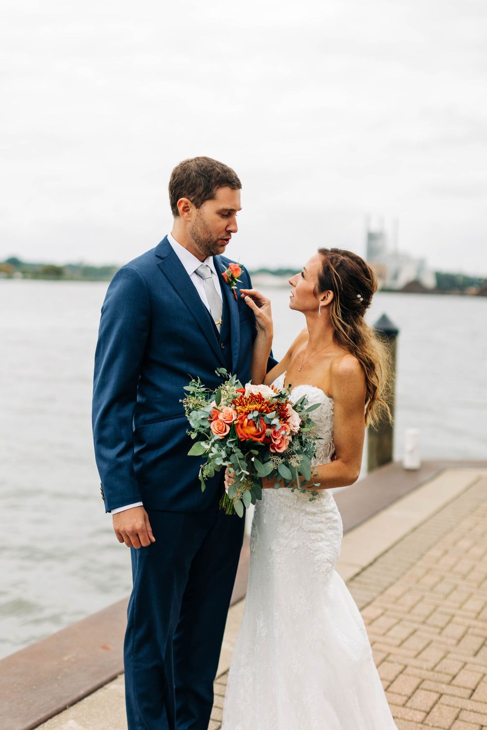 A couple in wedding attire shares a moment by the waterfront with a colorful bouquet.