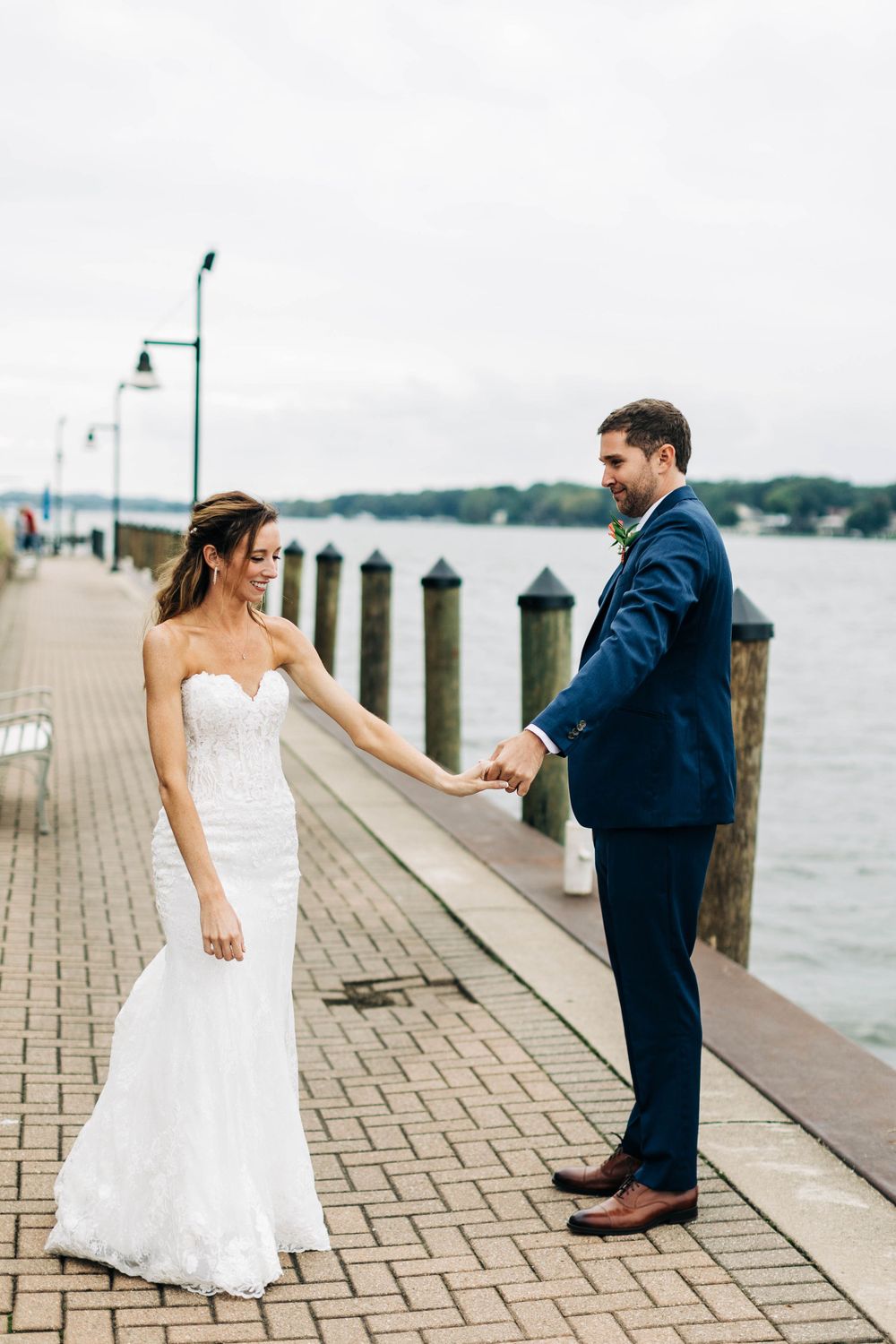 A couple in wedding attire holds hands on a wooden boardwalk by the water.