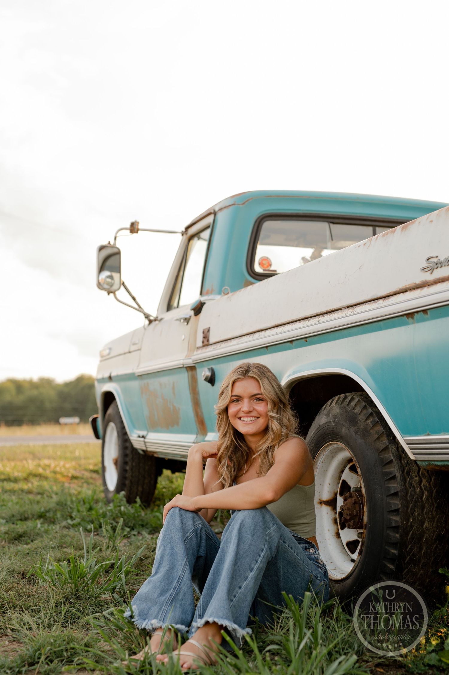 Someone poses beside a vintage turquoise pickup truck in a rural setting at sunset.