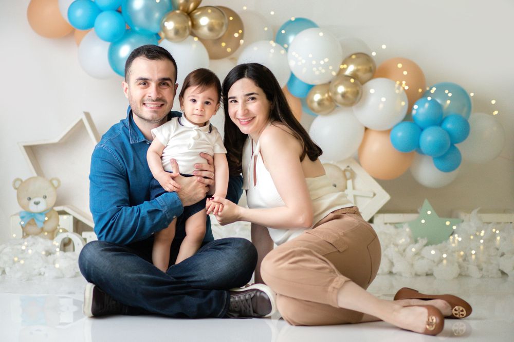 Family portrait with baby against blue and gold balloon arch backdrop in studio setting.