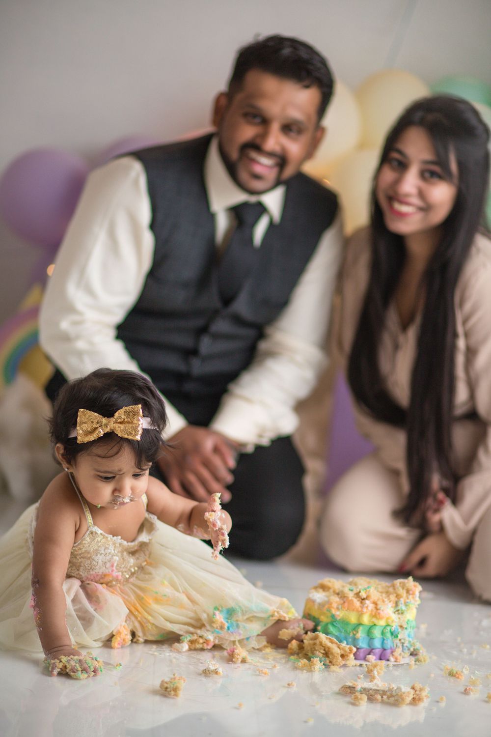 Baby in gold dress doing cake smash with colorful balloon background.