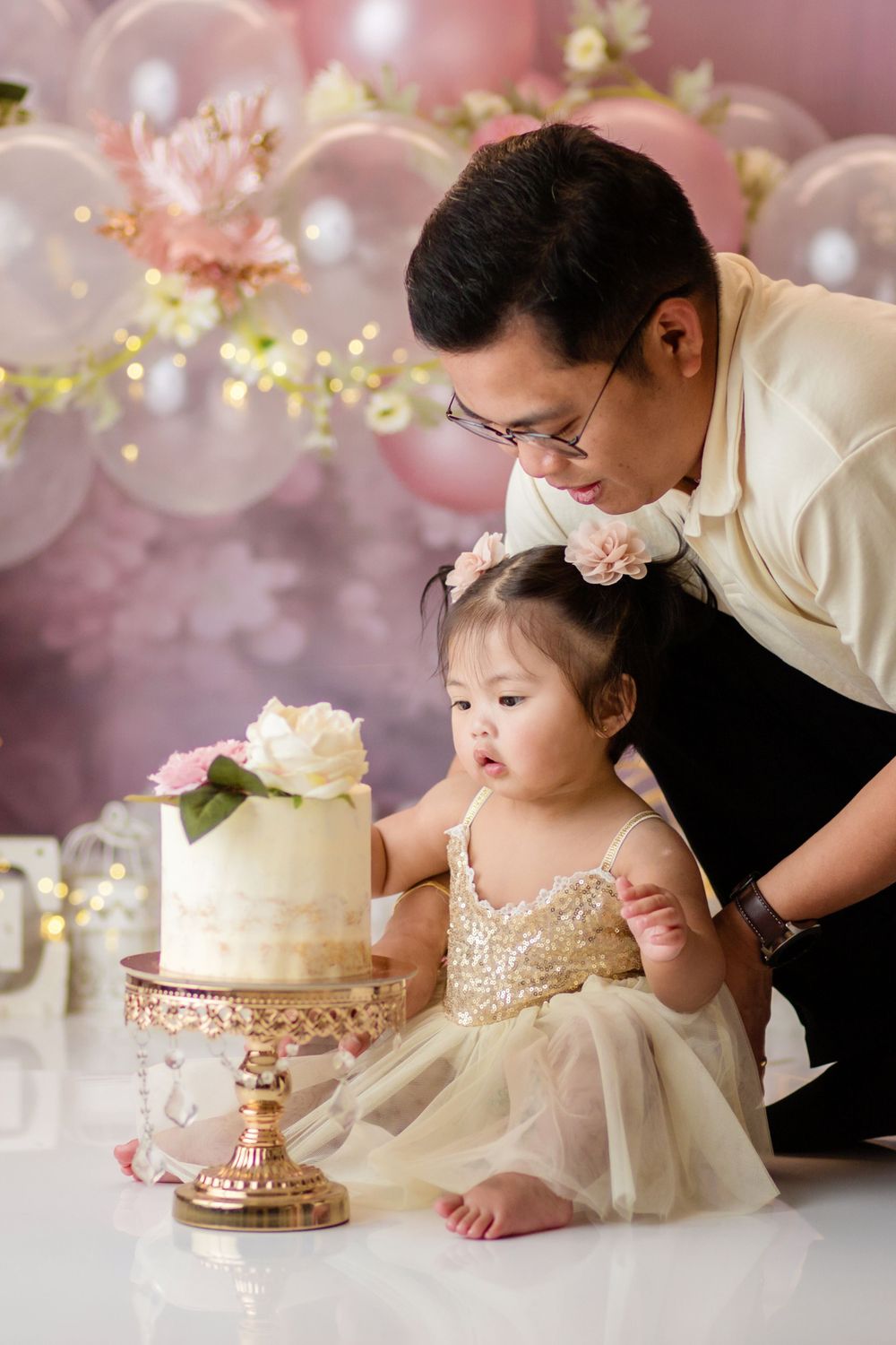 Baby in sparkly dress looking at elegant white cake with pink balloon decor background.