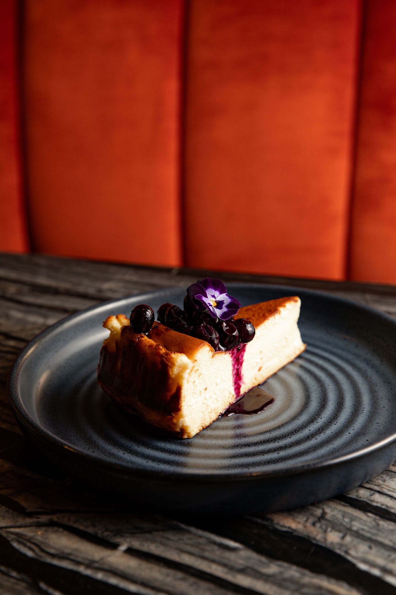 A slice of cheesecake with blueberry compote served on a black textured plate.