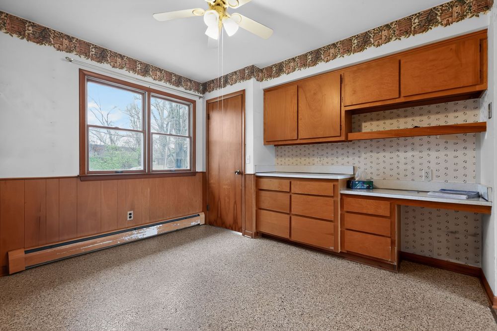 Vintage kitchen area with wood cabinets, built-in desk, baseboard heater, speckled floors, and wood-paneled walls.