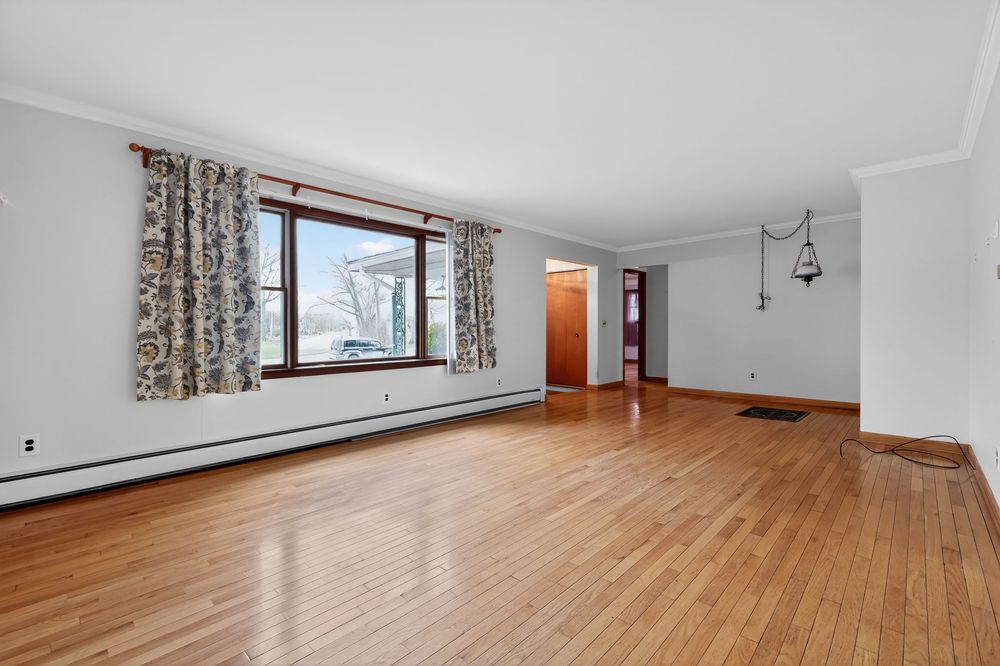 Bright living room with hardwood floors, large window with curtains, crown molding, and baseboard heating.