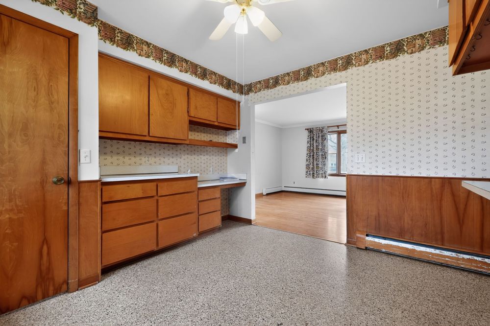 Vintage kitchen with wood cabinets, wallpaper border, speckled floor, and baseboard heater.