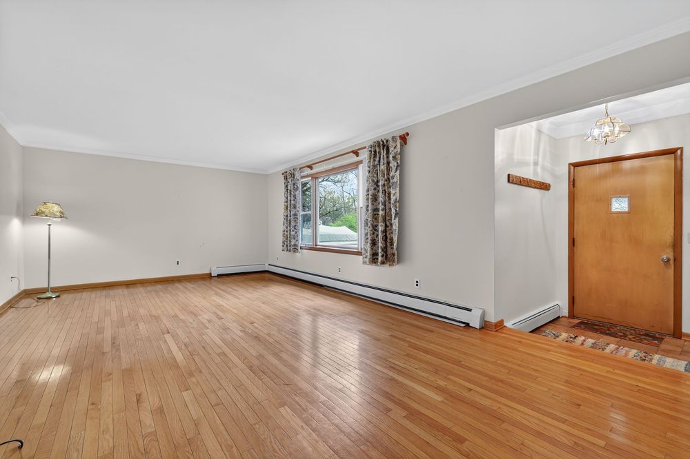 Bright living room with hardwood floors, baseboard heater, large window, and chandelier in entryway.