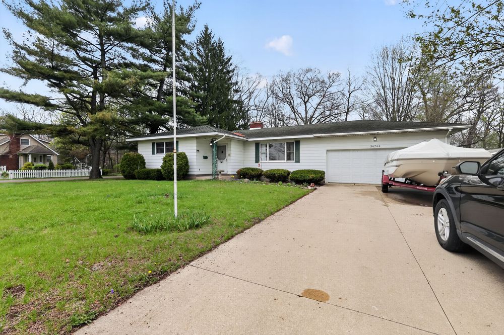 Ranch-style home with white exterior, attached garage, concrete driveway, and lush green lawn surrounded by mature trees.