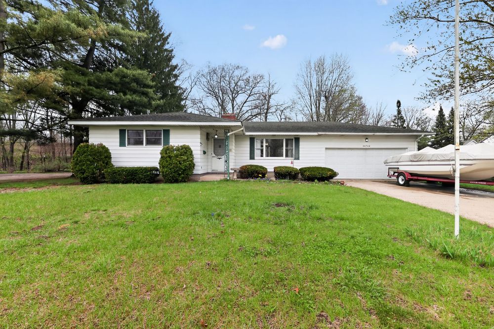 White ranch home with manicured lawn, attached garage, boat parked in driveway, and tall evergreen trees in background.