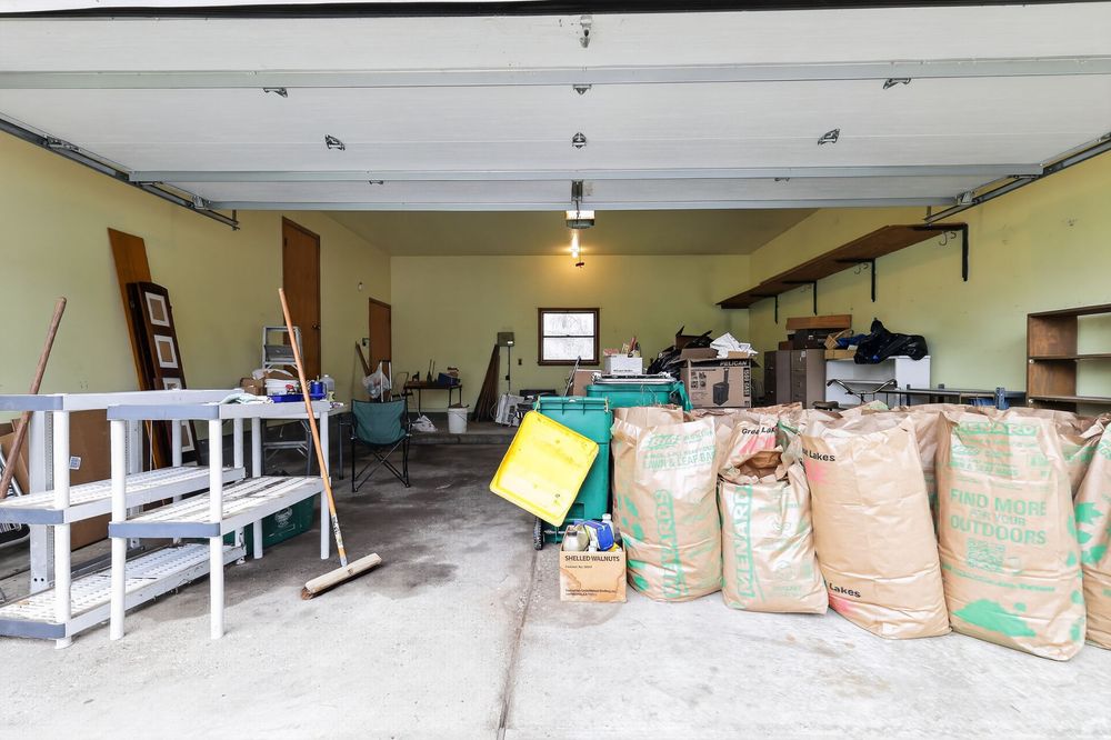 Open garage interior with shelving, stored bags of materials, and various tools and equipment on concrete floor.