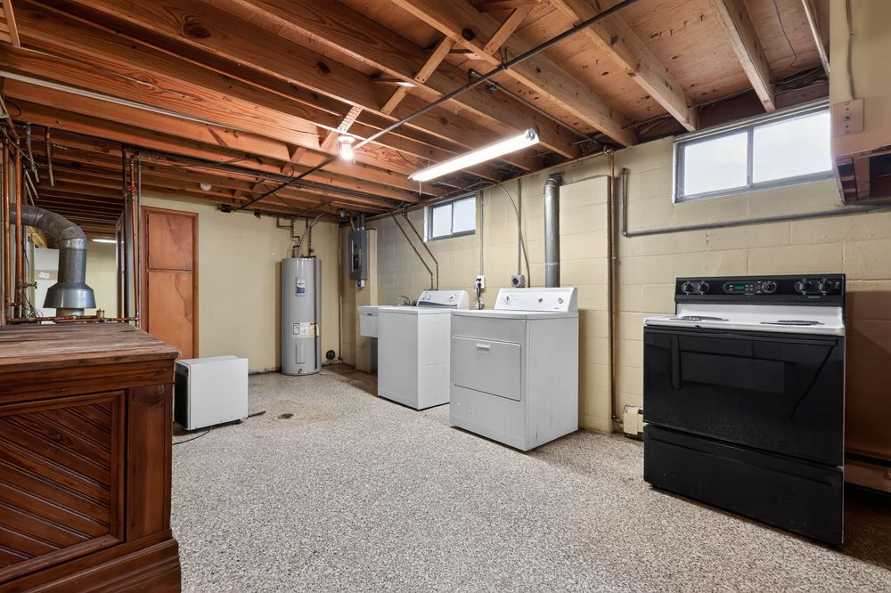 Basement laundry area with washer, dryer, black stove, water heater, and epoxy speckled flooring under exposed beams.