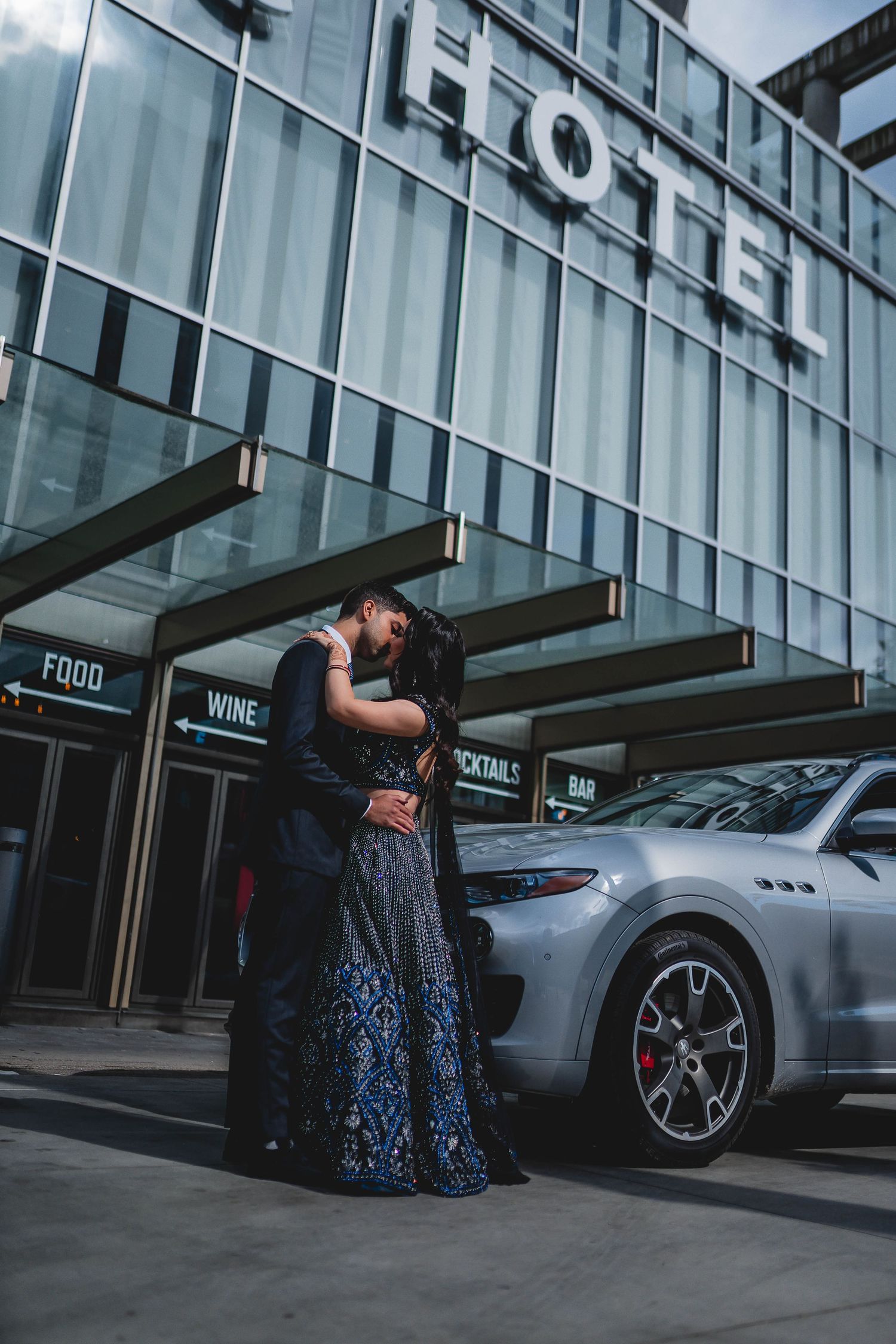 Romantic couple embraces outside modern hotel entrance with luxury silver car parked nearby.
