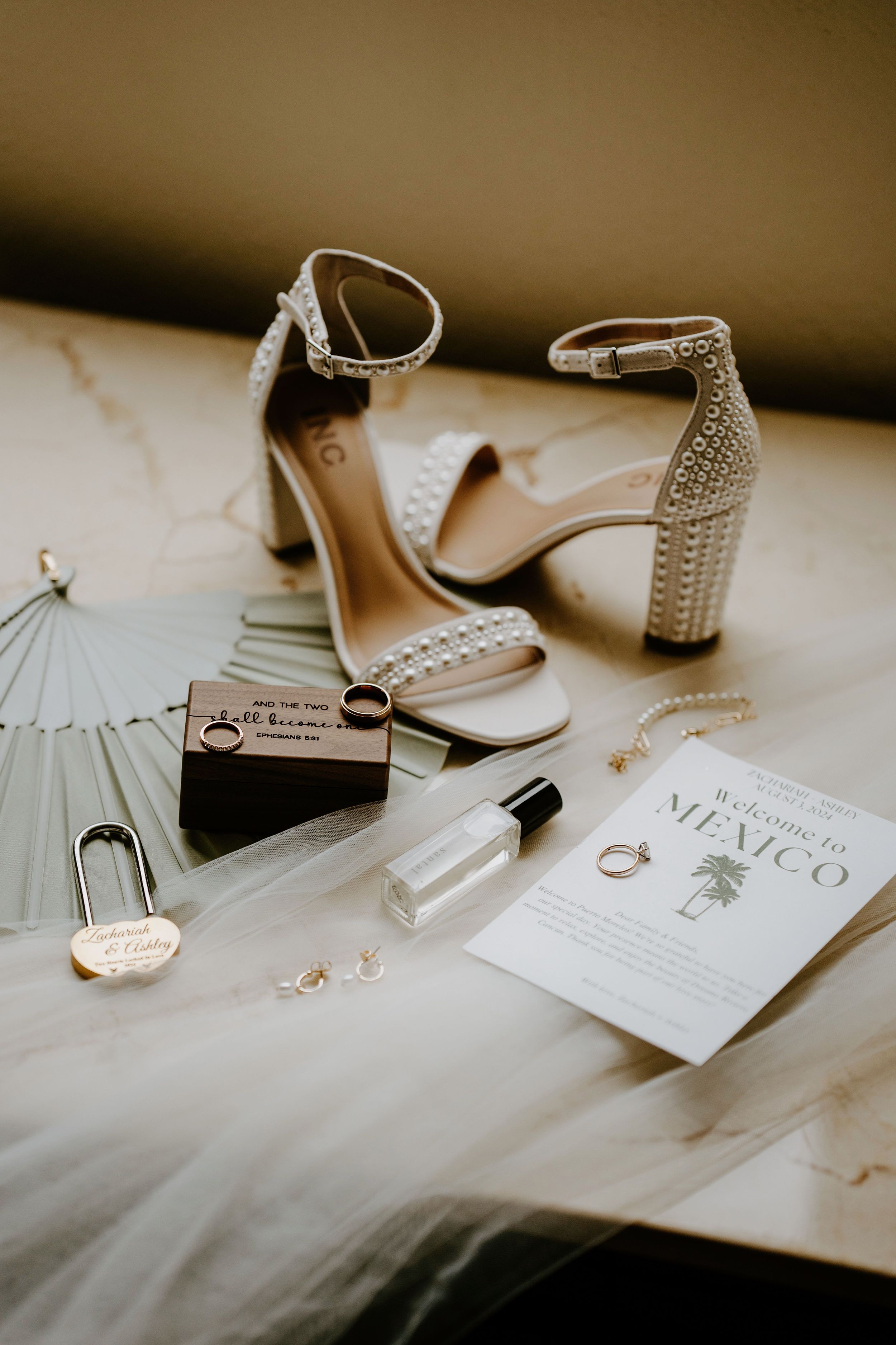 Wedding accessories including studded heels, perfume, rings and invitation laid out on white surface.