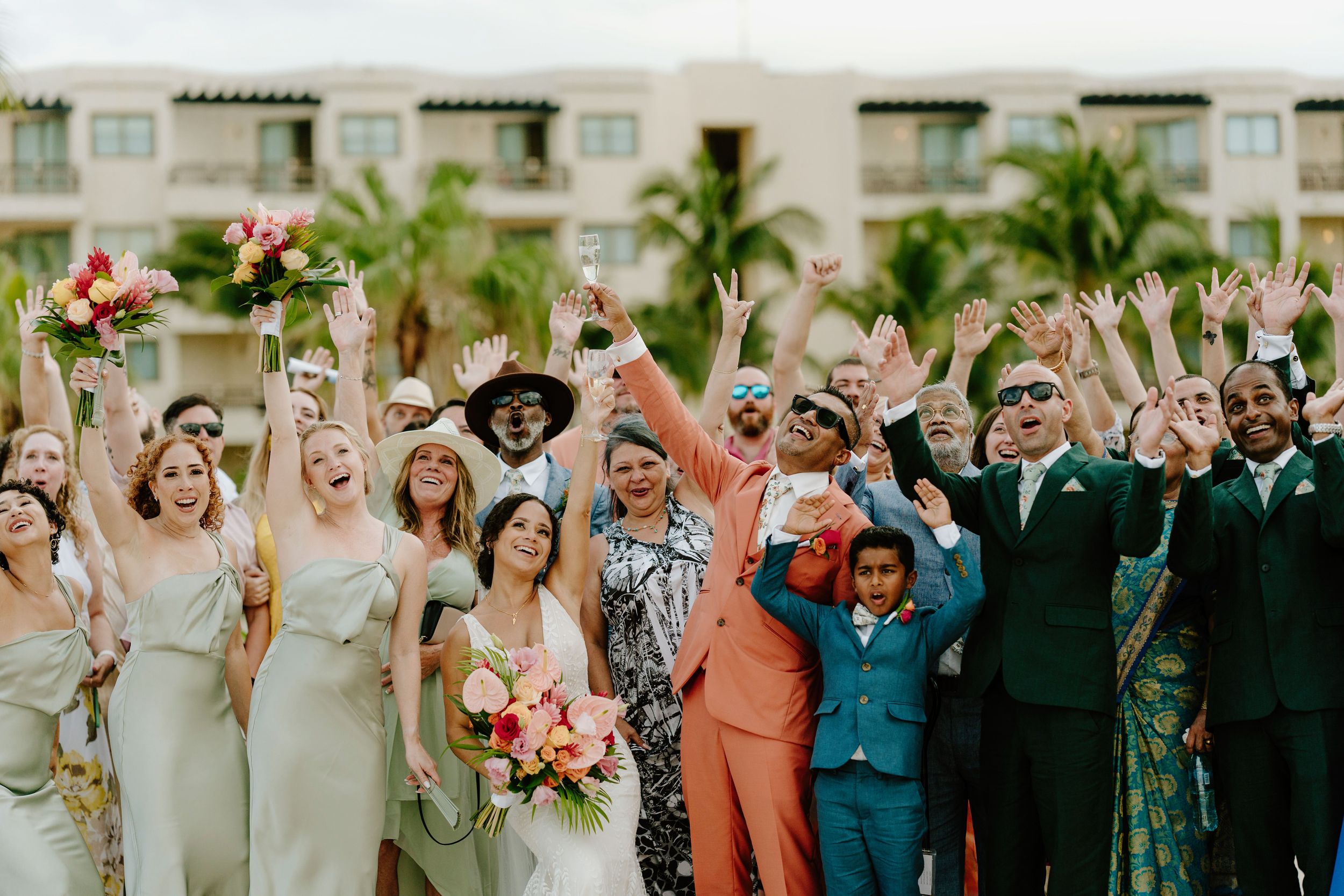 Wedding party celebrating with raised arms and colorful bouquets against resort backdrop.