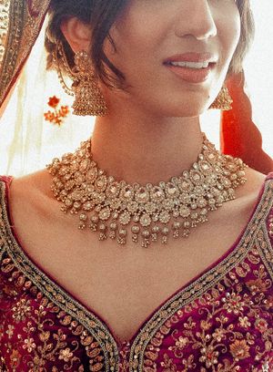 Close-up view of an ornate gold kundan necklace and matching earrings worn with a deep burgundy embroidered dress.