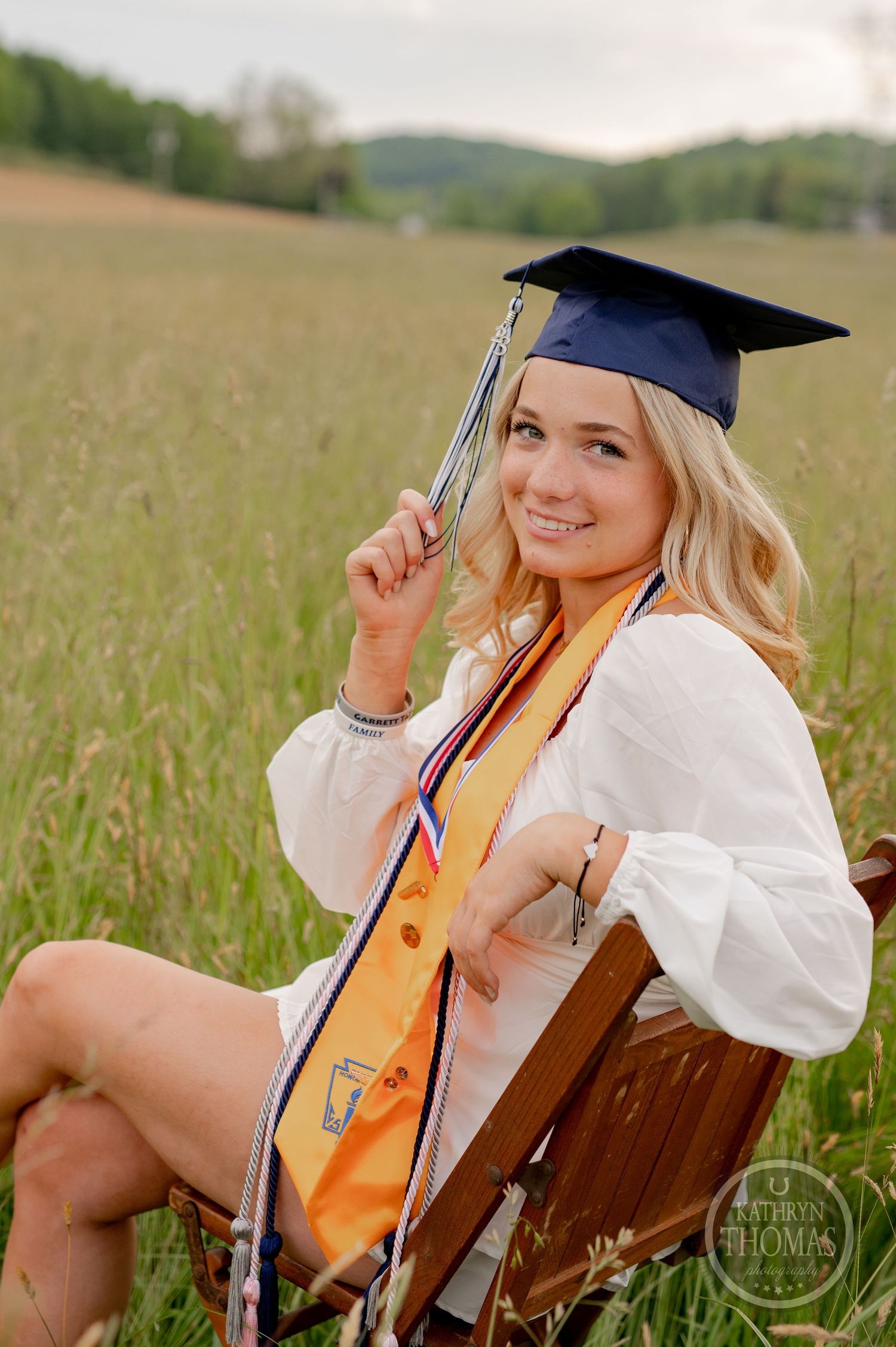 Graduate in cap with gold honors stole sits on vintage chair in rustic field setting.