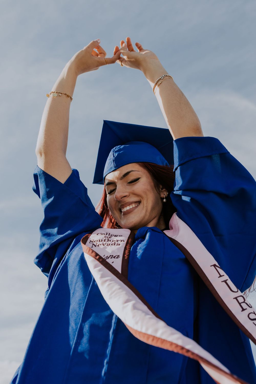 Smiling woman in blue graduation cap and gown raising arms at College of Southern Nevada ceremony.