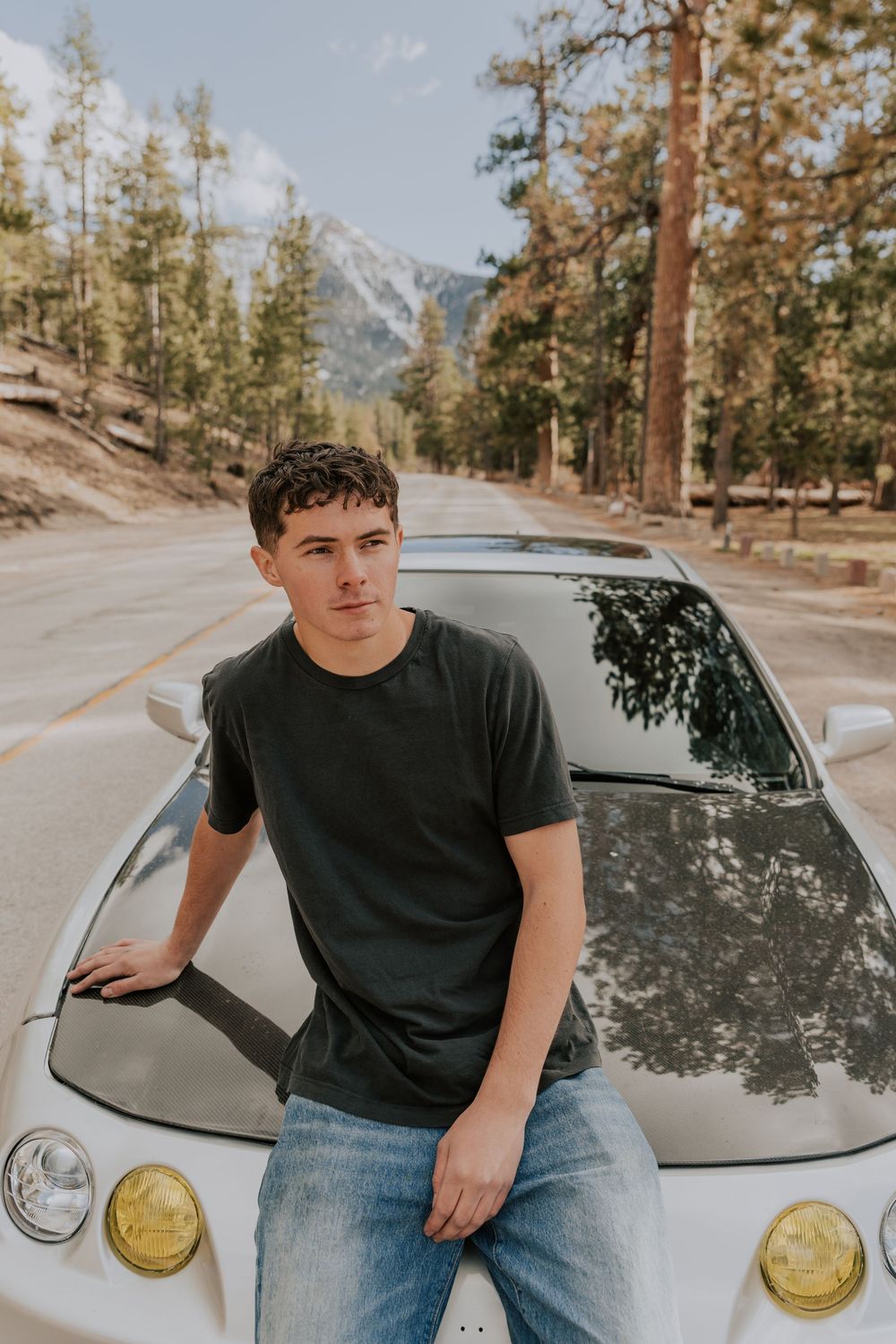 Teen boy leaning on silver sports car hood on mountain road with pine trees and peaks behind him.