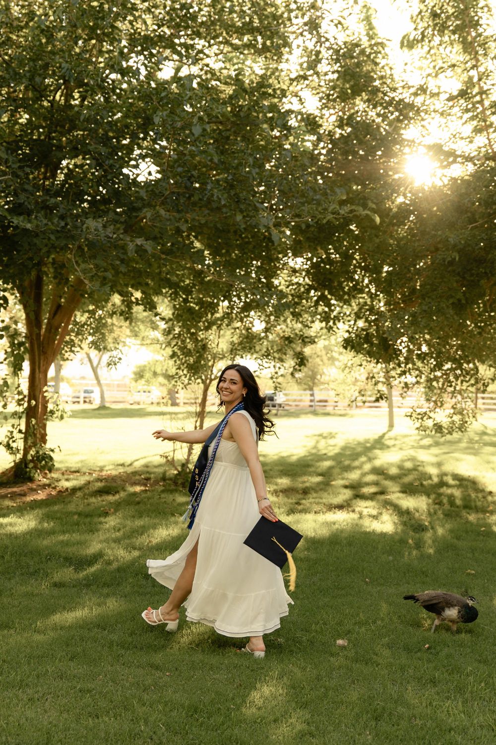 Smiling graduate in white dress holding cap walking on green grass with sunlight through trees.