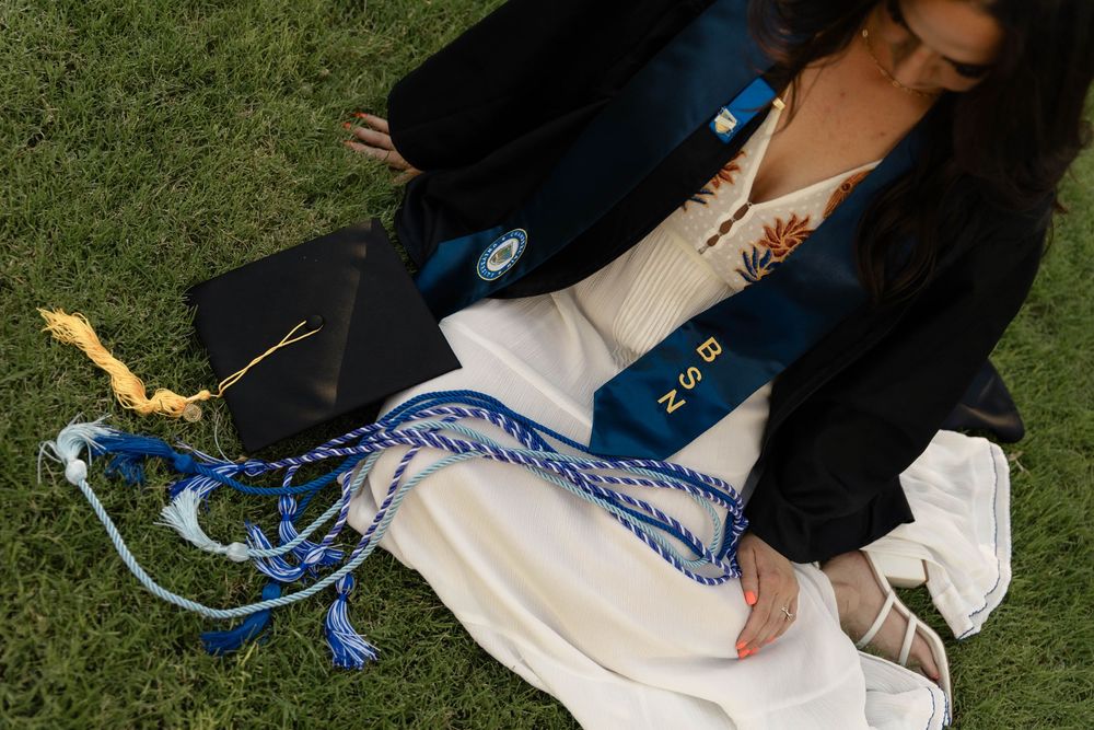 Close-up of graduate sitting on grass holding black cap with blue and white honor cords.