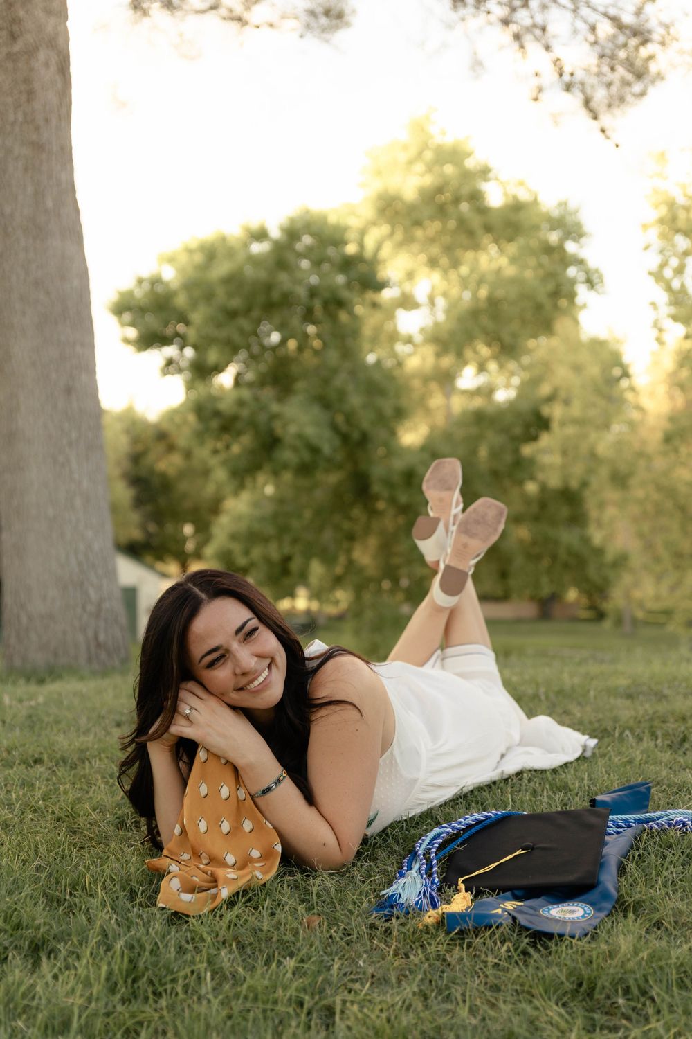 Smiling young woman in white dress lying on grass with graduation cap and honor cords nearby.