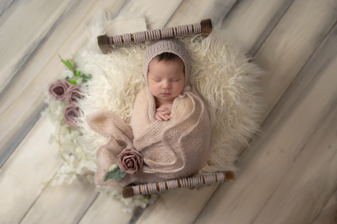 Newborn baby wrapped in blush pink swaddle with floral bonnet, sleeping peacefully in a fluffy white fur basket. Embracing Life’s Beauty.