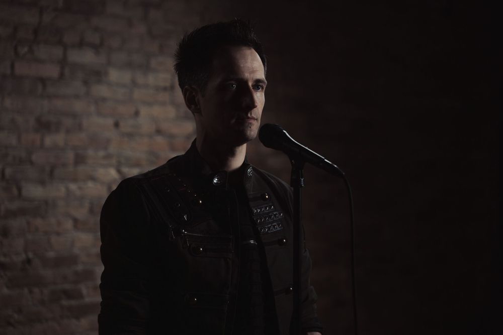 Silhouette of a performer standing at a microphone against a brick wall in moody lighting.