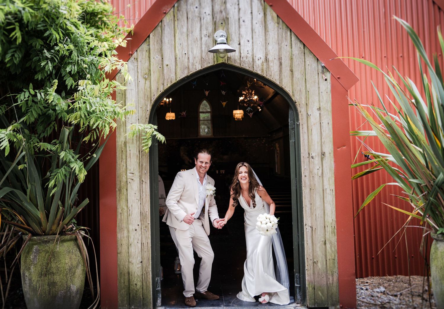 Newlyweds exiting rustic barn entrance with red walls and tropical plants.