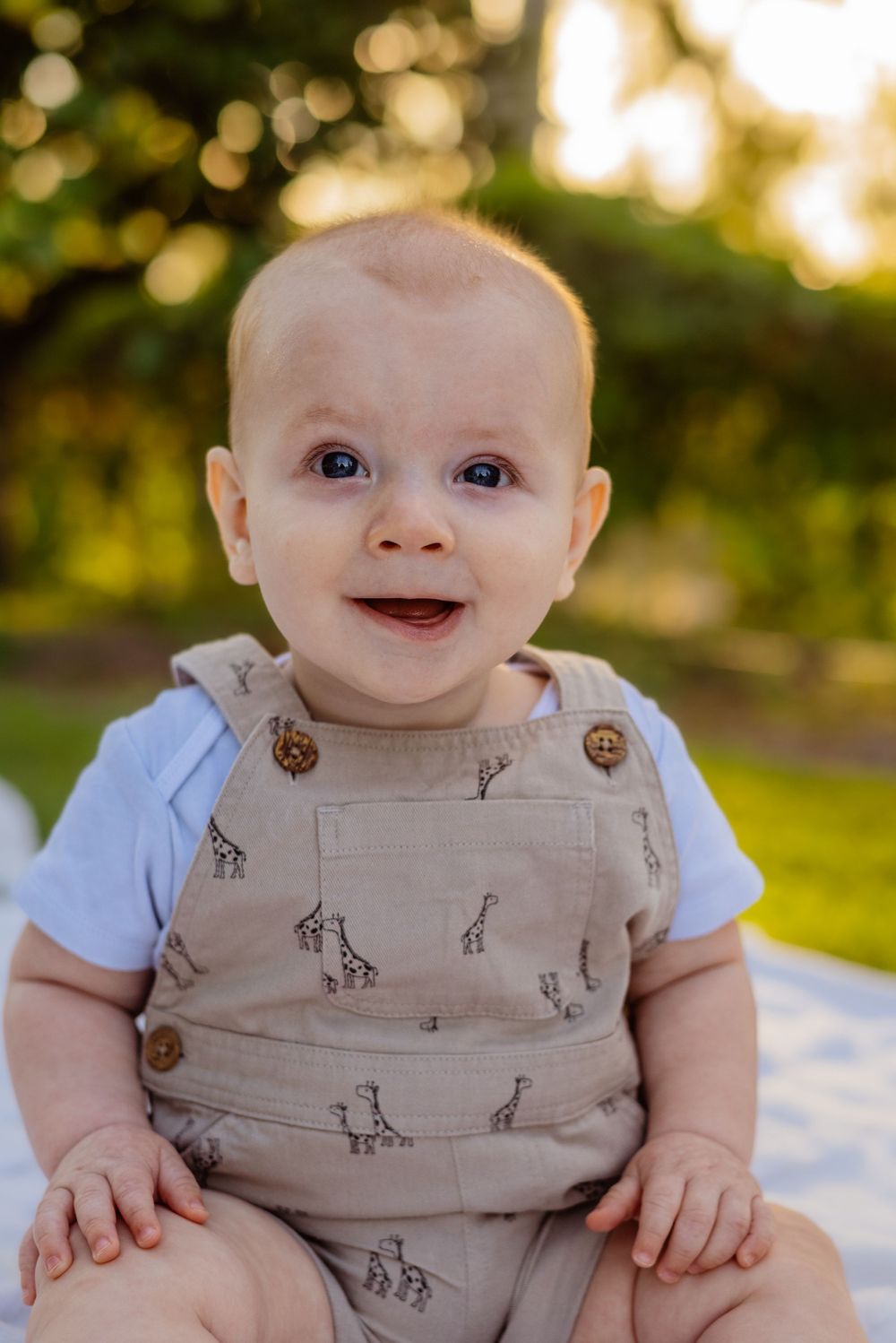 Baby wearing tan giraffe print overalls sits on white blanket in sunset garden setting.