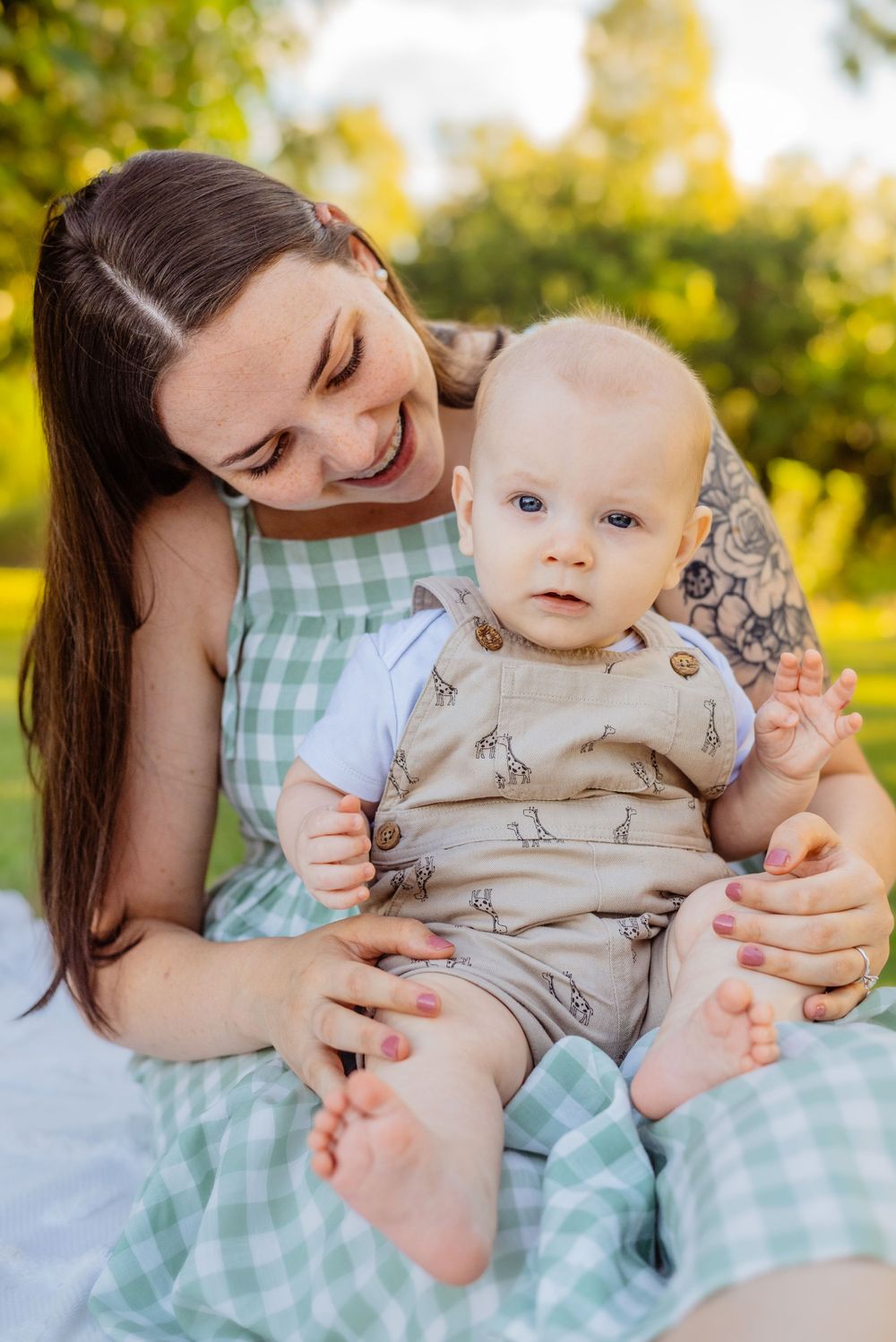 A baby in beige clothing sits on a green gingham dress in a sunny garden setting.