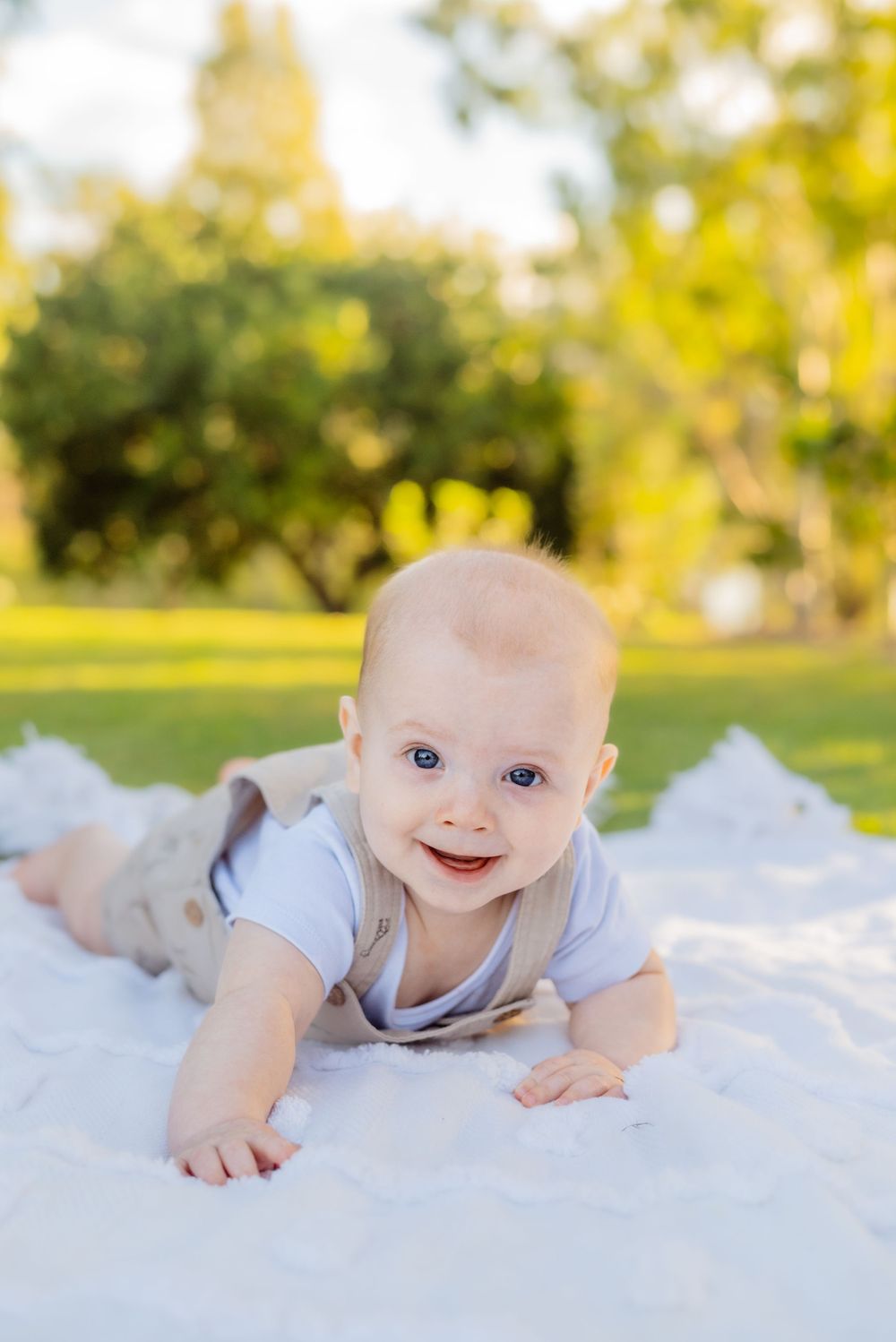 A smiling baby in light colored clothing crawls on a white blanket outdoors.