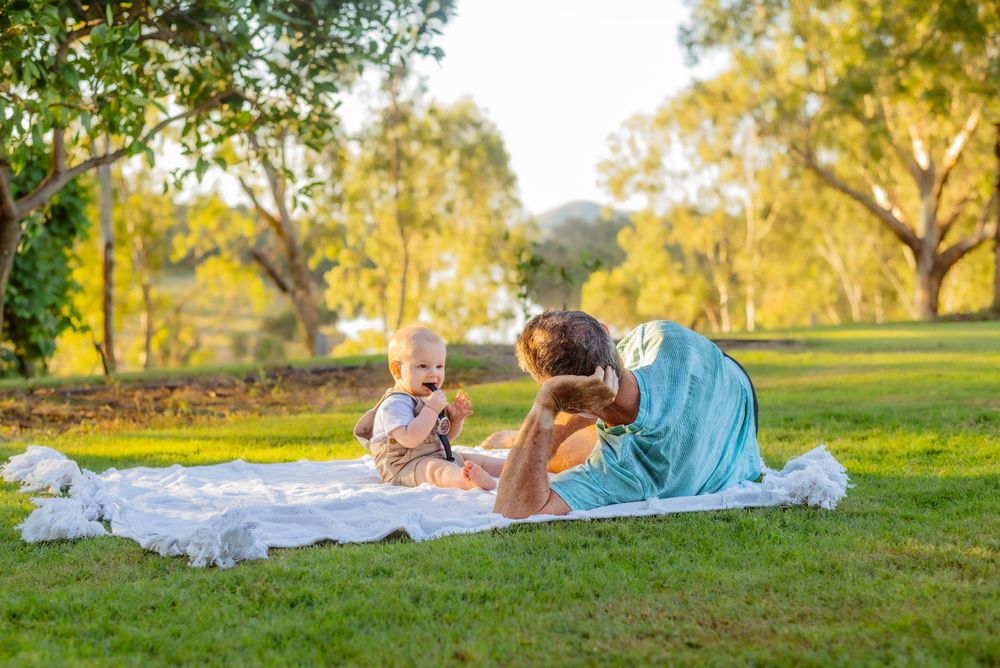 A family relaxes on a white blanket in a sunny park setting.